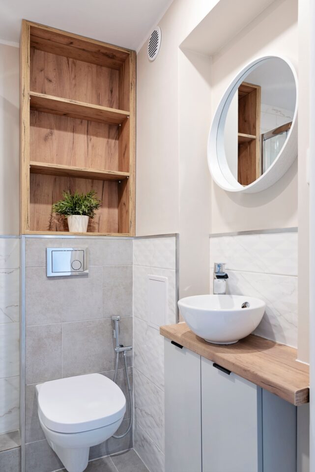 A small, compact bathroom remodeled to maximize space with a stone accent wall, vertical shelving unit, floating sink, and round mirror.